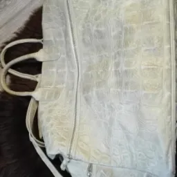 Używana, jasna torba damska ze wzorem imitującym skórę krokodyla, leżąca na brązowym futrze, widoczne zarysowania i ślady użytkowania.