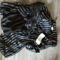 Czarna, cekinowa sukienka z papierową metką leżąca na drewnianej podłodze.