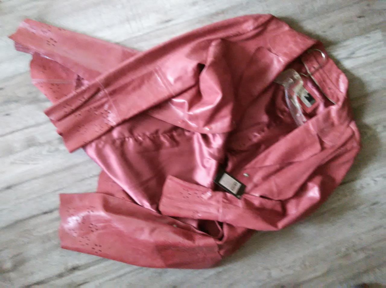 Różowa, błyszcząca kurtka z ekoskóry z ażurowymi detalami na mankietach, leżąca na podłodze z widoczną metką i wieszakiem.