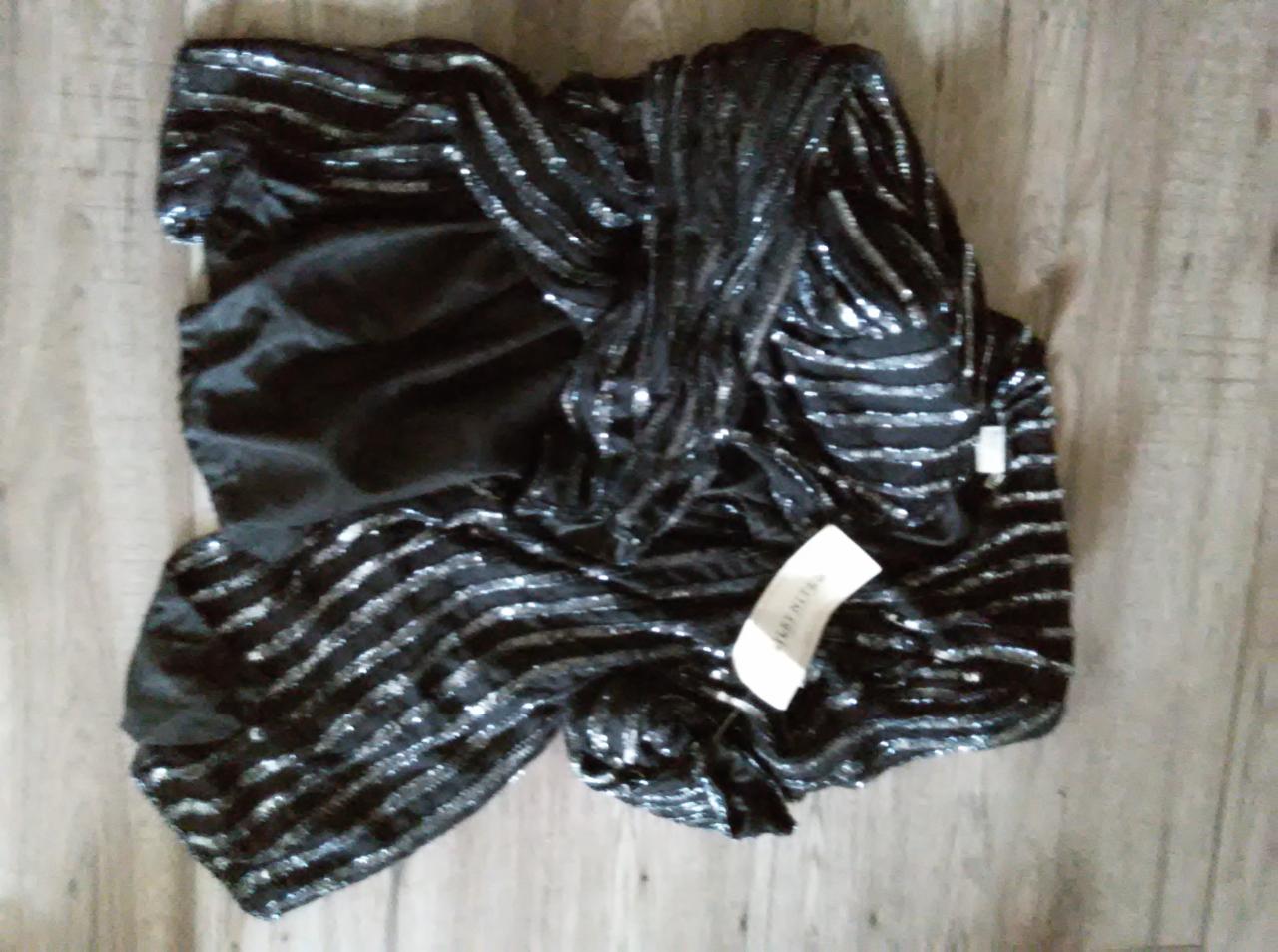 Czarna, cekinowa sukienka z papierową metką leżąca na drewnianej podłodze.