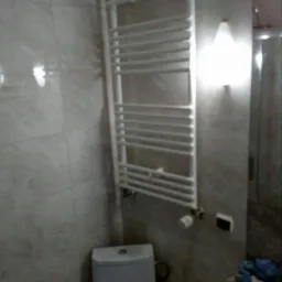 Biały, drabinkowy grzejnik łazienkowy zamontowany na ścianie z płytek imitujących marmur, obok toaleta i częściowo widoczna umywalka, oświetlenie punktowe nad grzejnikiem.