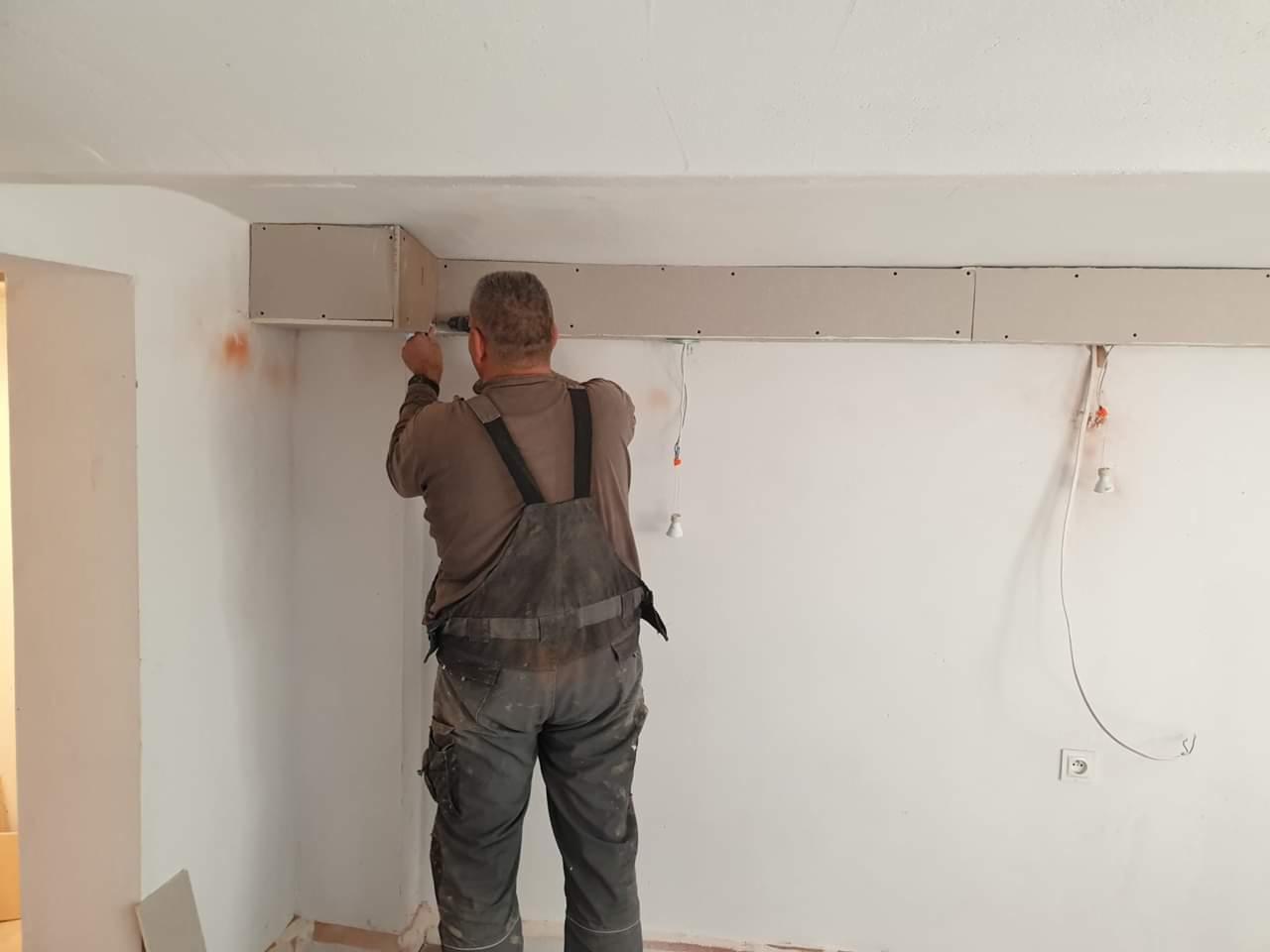 Pracownik w kombinezonie montuje konstrukcję z płyt gipsowo-kartonowych przy suficie, widoczne przewody elektryczne i fragmenty instalacji.