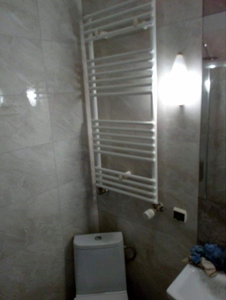 Biały, drabinkowy grzejnik łazienkowy zamontowany na ścianie z płytek imitujących marmur, obok toaleta i częściowo widoczna umywalka, oświetlenie punktowe nad grzejnikiem.