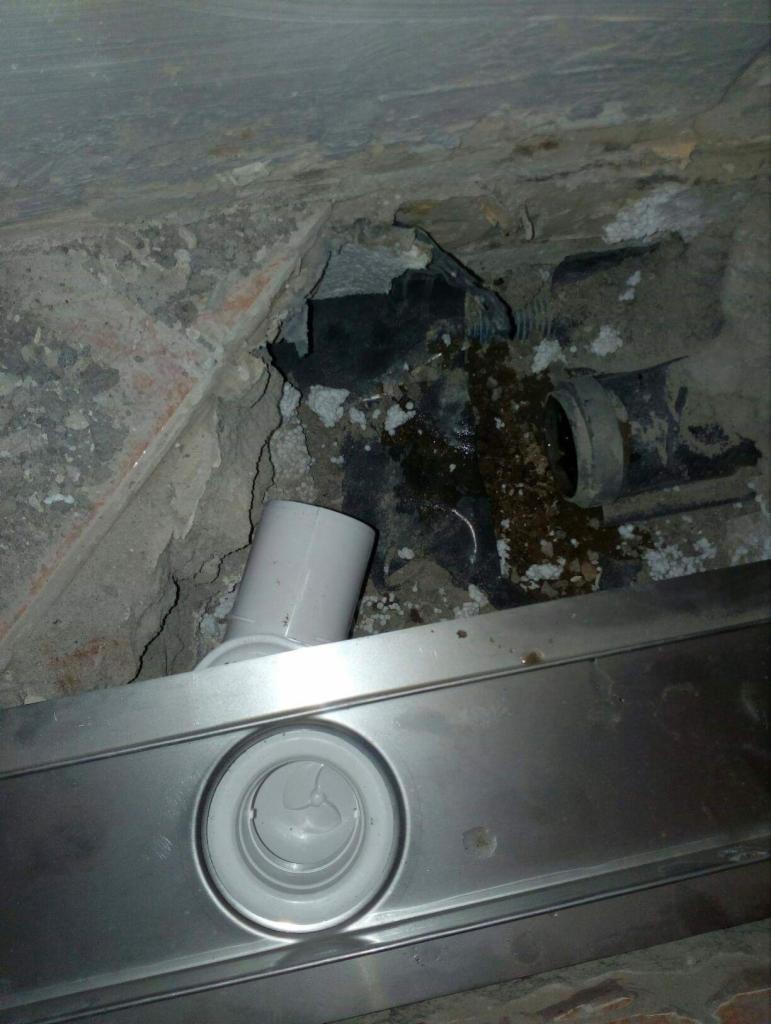 Nieszczelny odpływ liniowy w surowym stanie, widoczne zacieki i zabrudzenia na betonowym podłożu, czarna rura kanalizacyjna z widocznymi śladami wycieków.