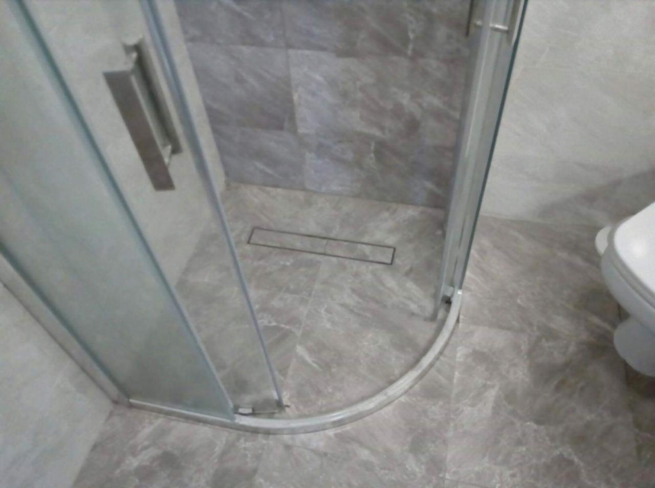 Szklana kabina prysznicowa z odpływem liniowym w posadzce, wykończona płytkami imitującymi marmur w odcieniach szarości, z widocznym fragmentem toalety.