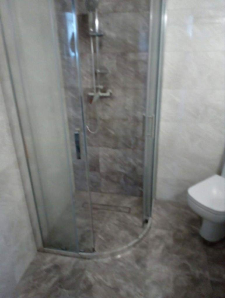 Szara łazienka z kabiną prysznicową z przezroczystego szkła i odpływem liniowym w podłodze z marmuru, widoczna również toaleta.