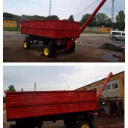 Czerwona przyczepa rolnicza z żółtymi kołami i podajnikiem ślimakowym, stojąca na placu przed budynkiem przemysłowym.