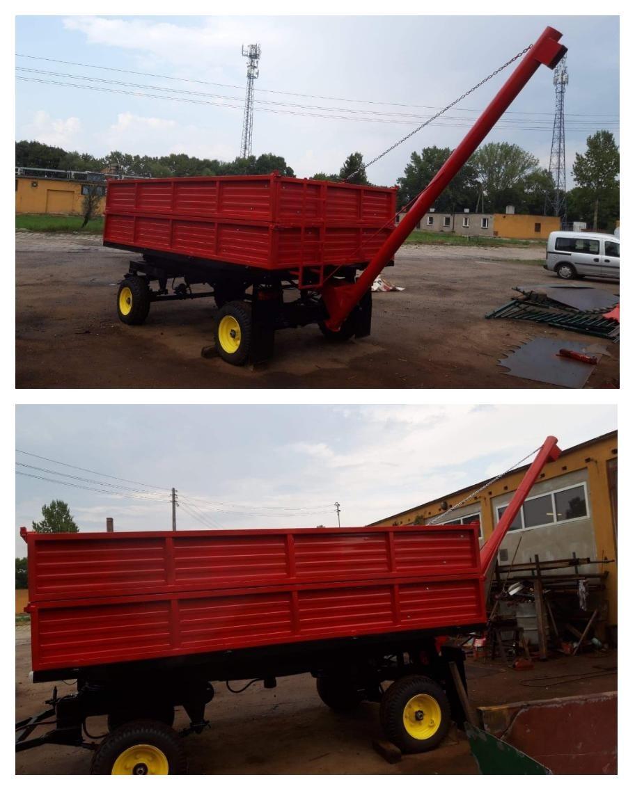 Czerwona przyczepa rolnicza z żółtymi kołami i podajnikiem ślimakowym, stojąca na placu przed budynkiem przemysłowym.