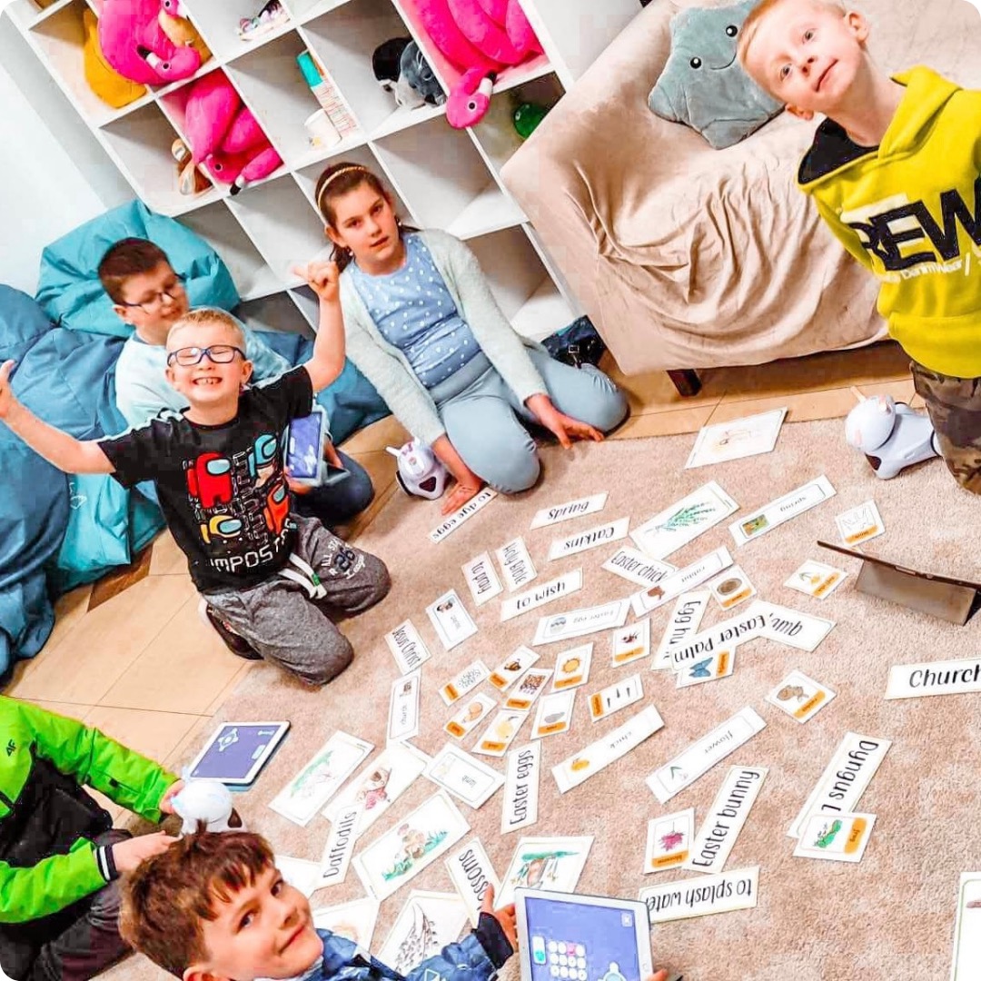 Grupa dzieci siedzi na podłodze wokół rozłożonych kart obrazkowych z angielskimi słowami związanymi z Wielkanocą i wiosną, używając tabletów i interaktywnych robotów do nauki.