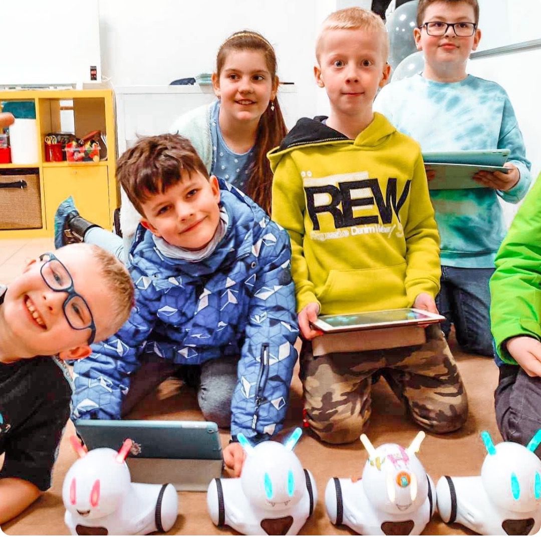 Grupa dzieci w różnym wieku siedzi na podłodze z tabletami, otoczona zabawkami-robotami z podświetlanymi uszami, w tle regał z zabawkami.