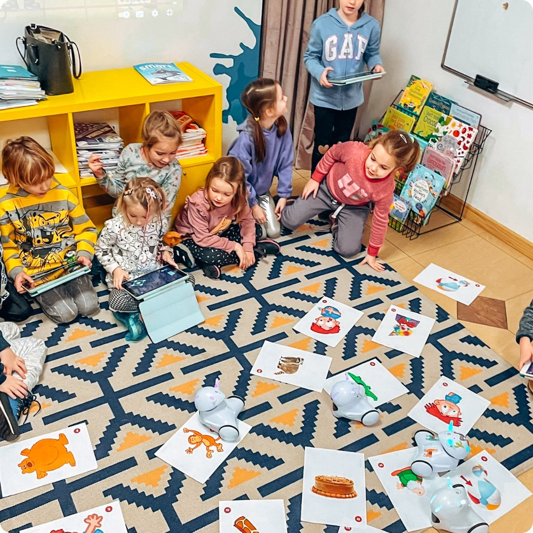 Grupa dzieci siedzi na dywanie w klasie, używając tabletów i interaktywnych robotów do nauki, z żółtym regałem i książkami w tle.