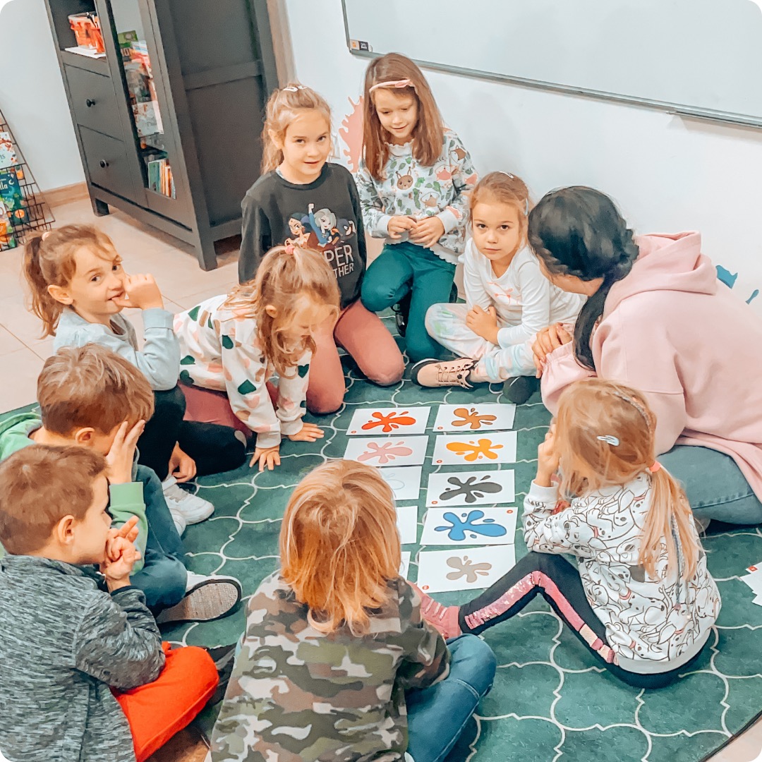 Grupa dzieci siedzi na podłodze i gra w grę edukacyjną z kolorowymi plamami, nadzorowana przez kobietę w różowej bluzie.