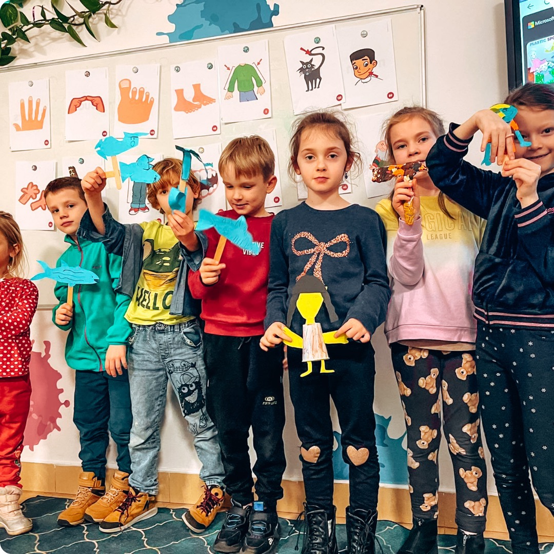 Grupa dzieci prezentuje prace plastyczne: papierowe ryby na patykach i postać z papieru, na ścianie ilustracje części ciała z podpisami.