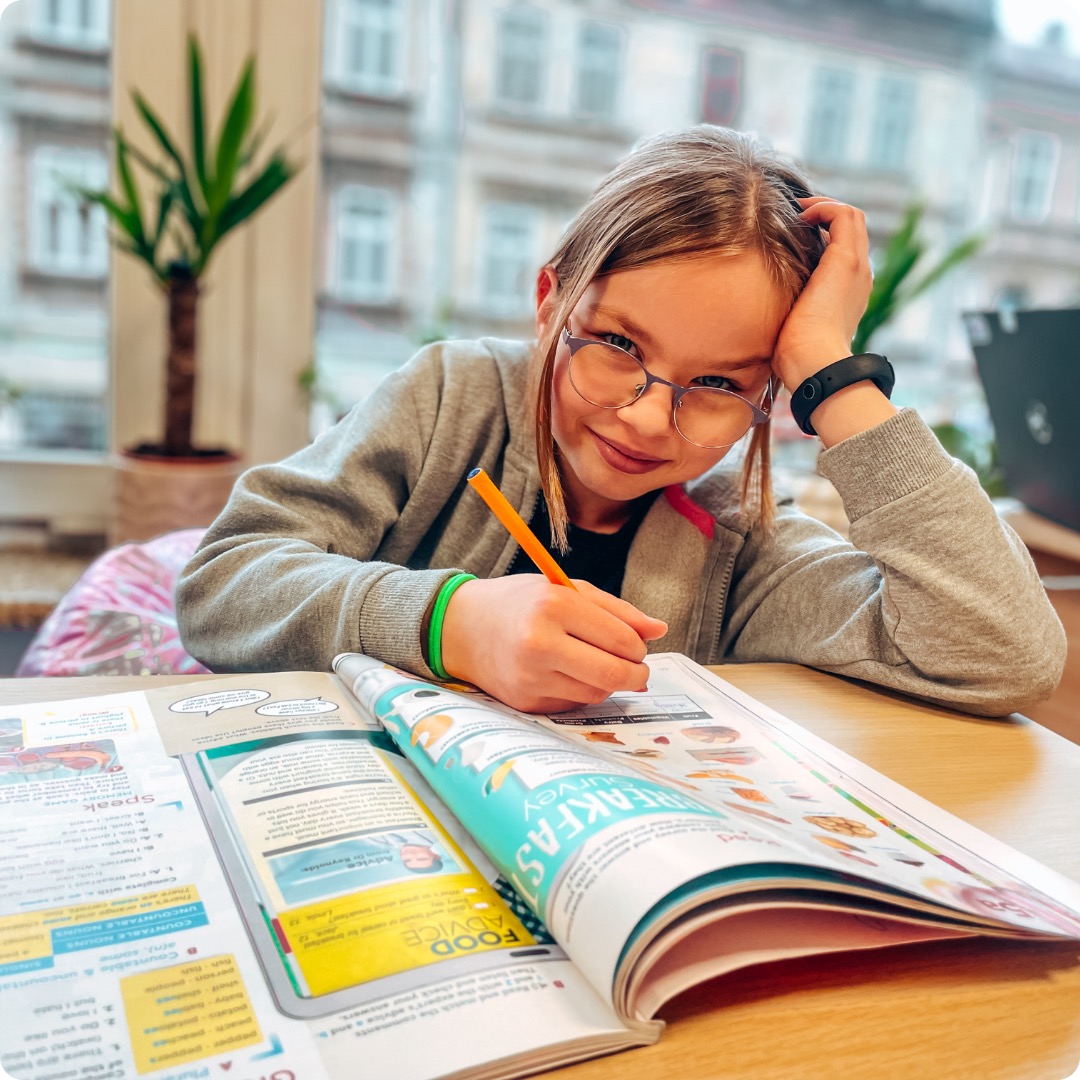 Ucząca się dziewczynka w okularach rozwiązuje ćwiczenia z angielskiego, używając pomarańczowego ołówka i podręcznika z poradami dotyczącymi śniadania.