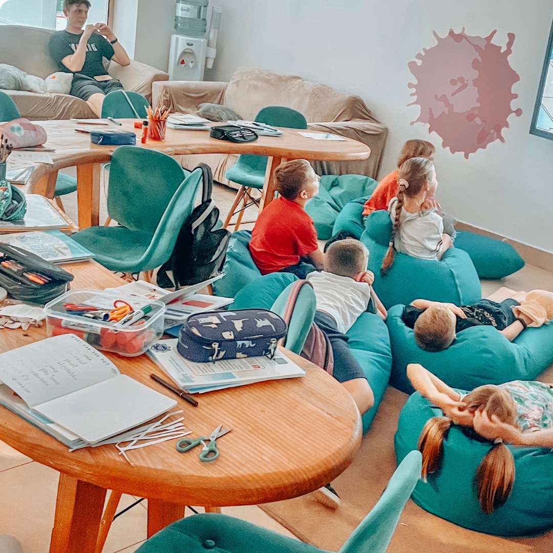 Grupa dzieci siedzi na turkusowych pufach w sali lekcyjnej, z drewnianymi stolikami pełnymi przyborów szkolnych i nauczycielem w tle.