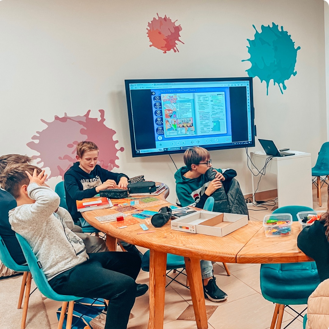 Uczniowie przy okrągłym stole podczas lekcji z interaktywną tablicą, na ścianie kolorowe plamy, widoczne podręczniki i akcesoria szkolne.