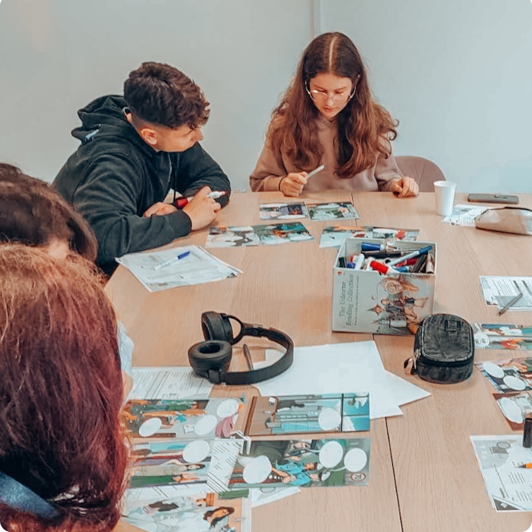 Uczniowie pracują z kartami obrazkowymi i markerami przy stole podczas zajęć językowych, słuchawki leżą na stole.