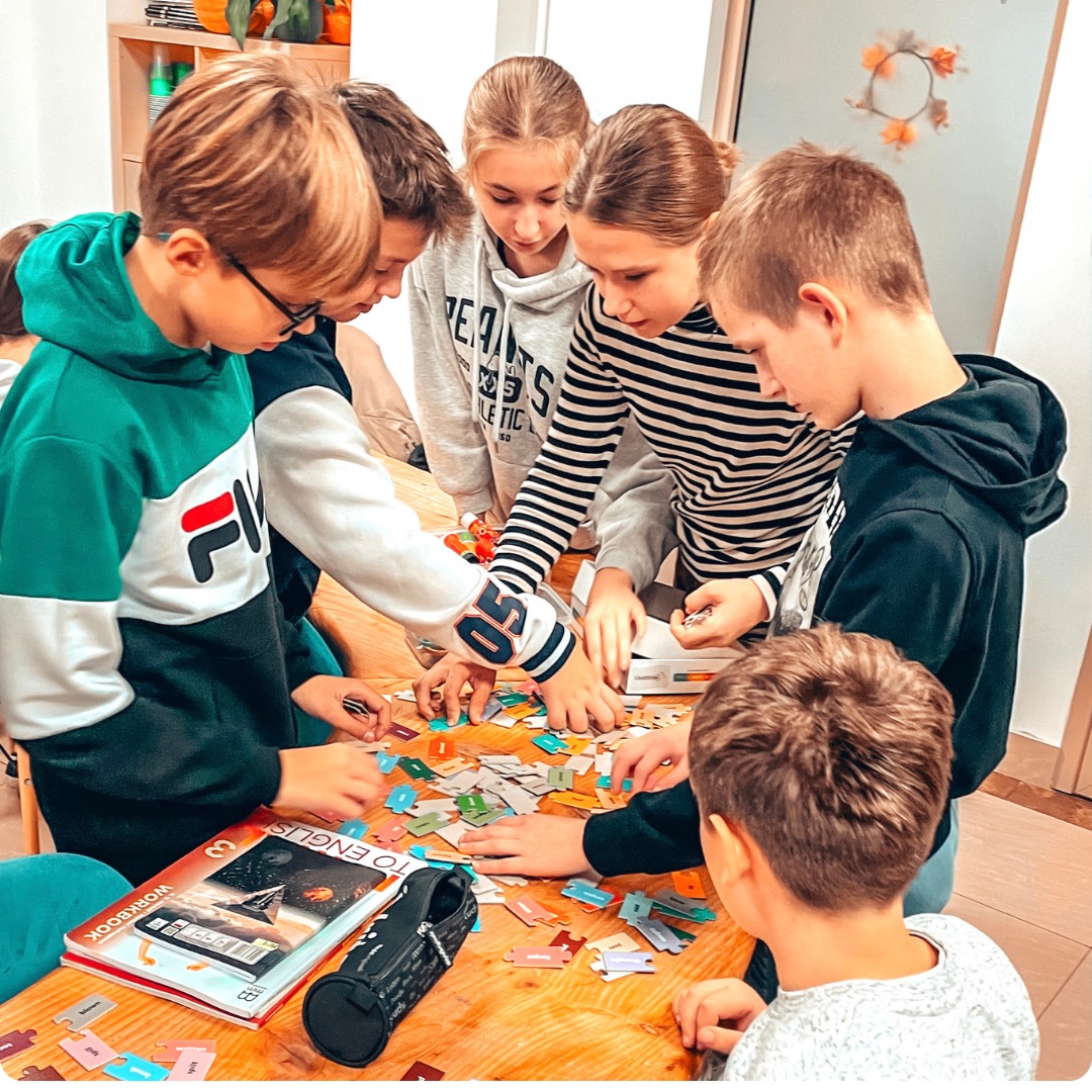Grupa uczniów skupiona nad drewnianym stołem, układająca kolorowe puzzle językowe, obok leży zeszyt do angielskiego i piórnik.