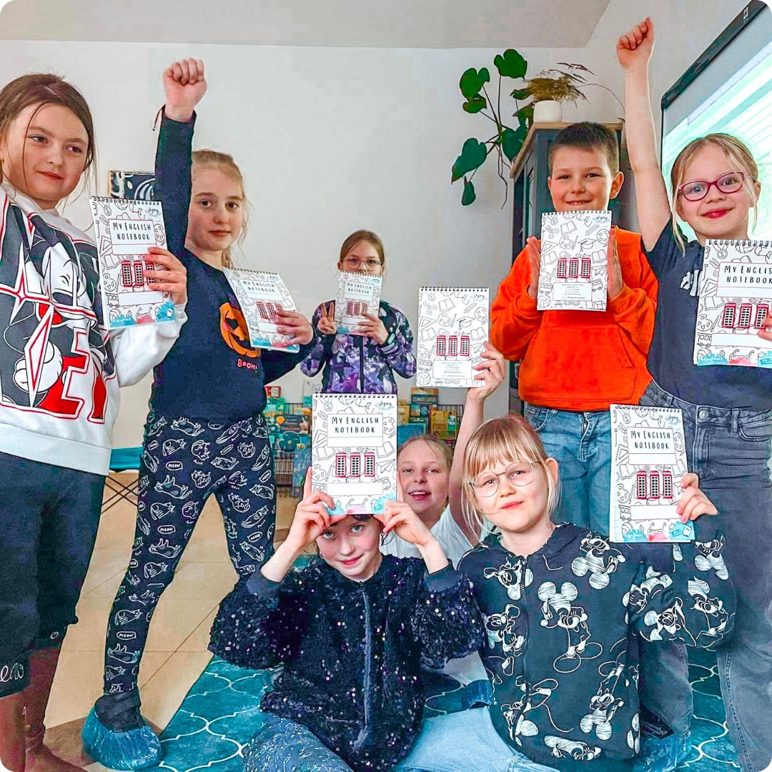 Grupa uśmiechniętych dzieci prezentuje zeszyty do angielskiego z motywem budki telefonicznej, część z podniesionymi rękami w geście radości.
