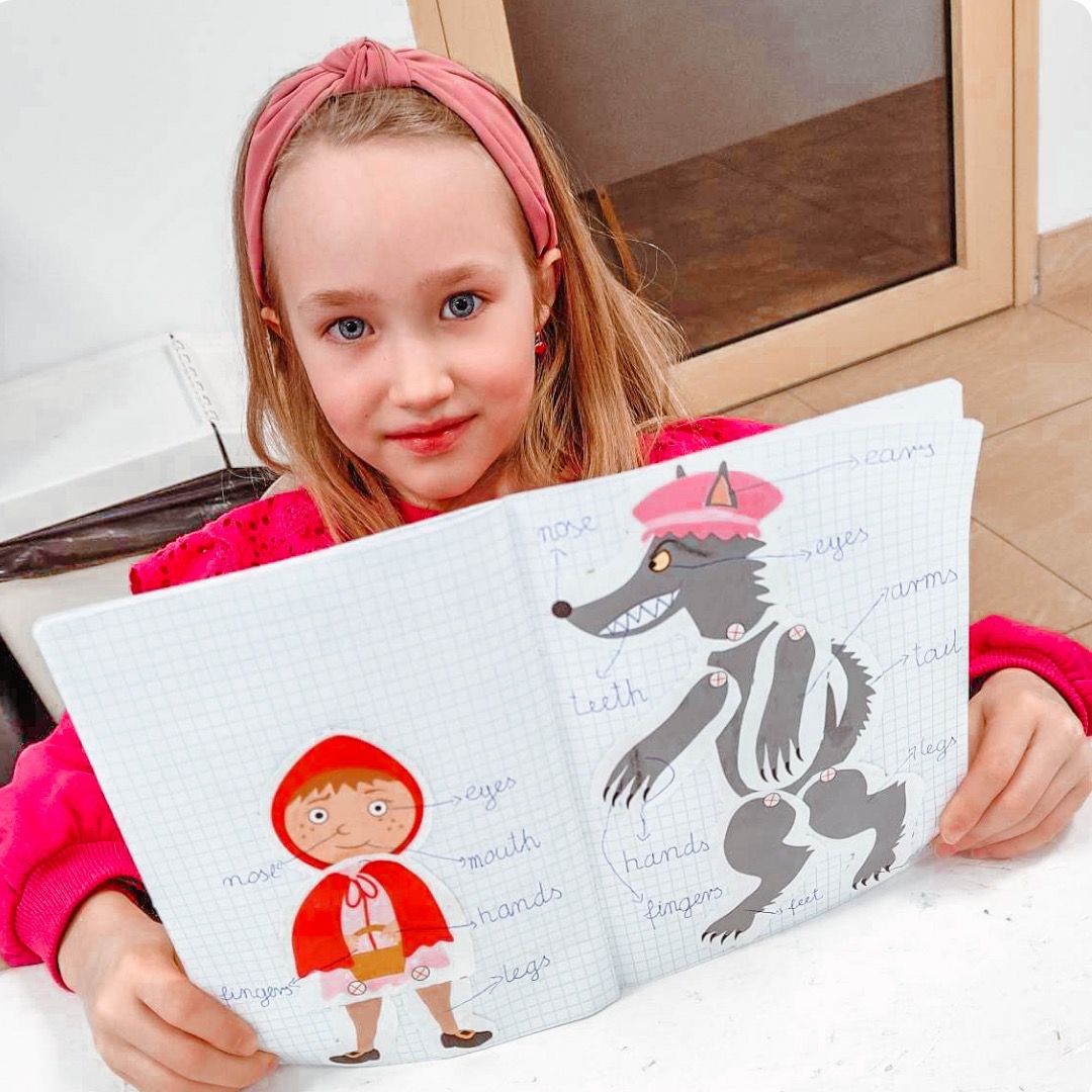 Dziewczynka z różową opaską prezentuje otwarty zeszyt w kratkę z ilustracjami Czerwonego Kapturka i wilka z podpisami części ciała.