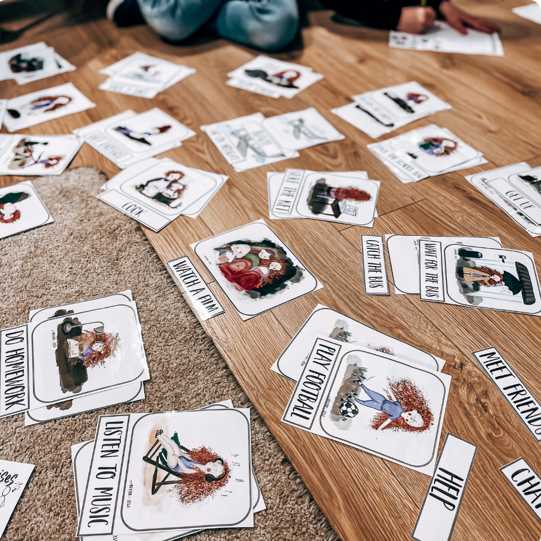 Karty edukacyjne z ilustracjami i podpisami czynności, rozłożone na drewnianej podłodze i dywanie, używane do nauki języka angielskiego.