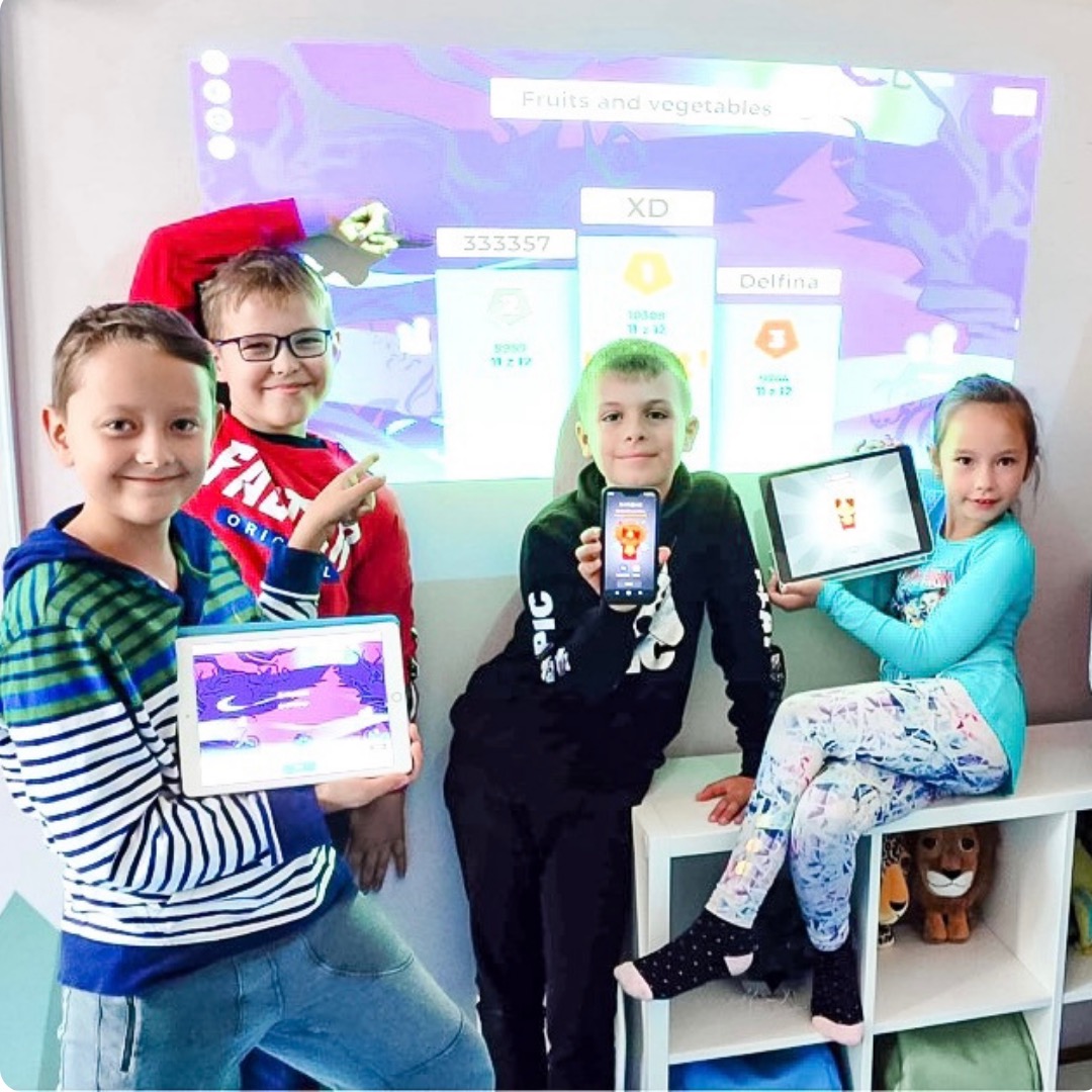 Grupa dzieci prezentuje interaktywną lekcję o owocach i warzywach na tabletach i rzutniku, z wyświetlonymi wynikami i rankingiem.