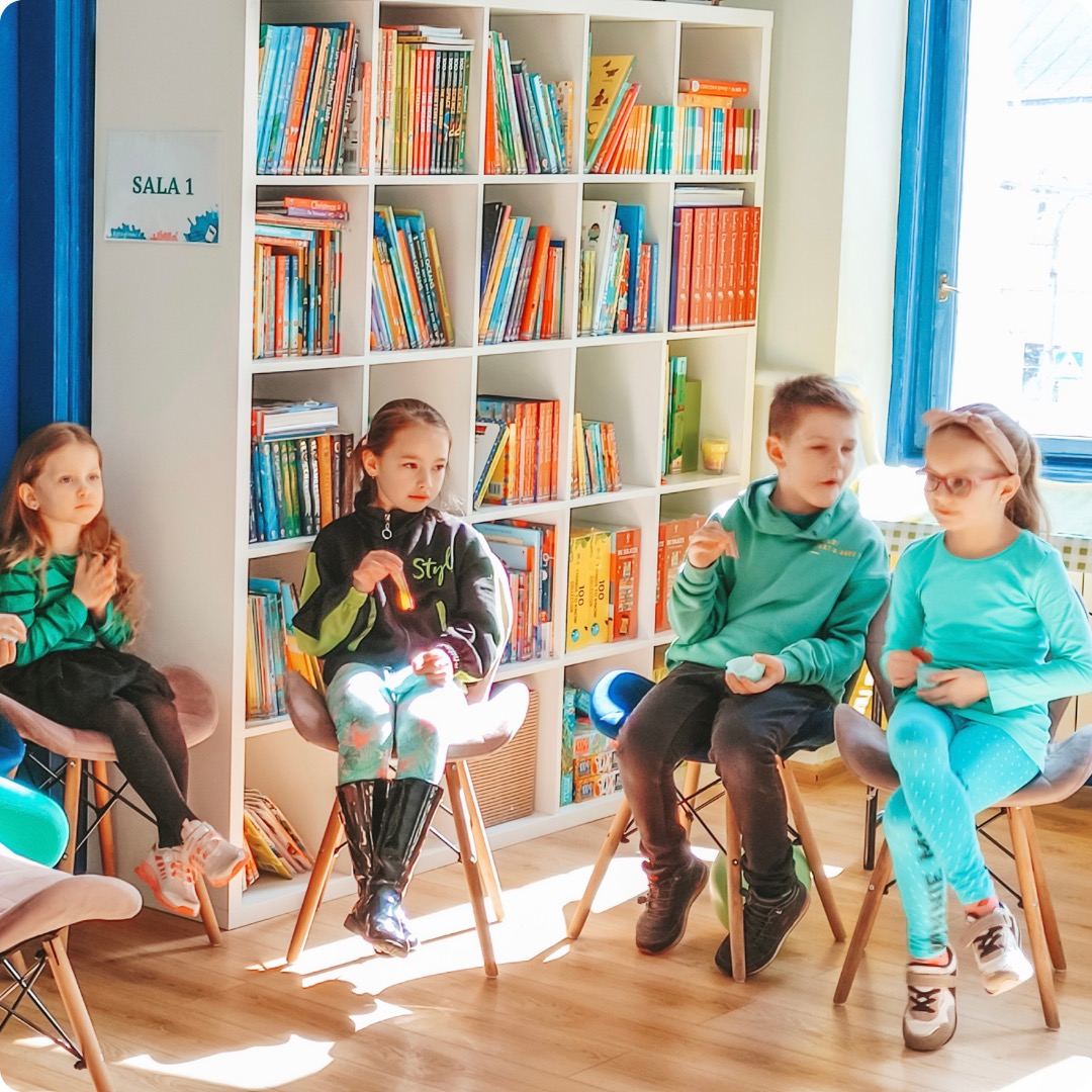 Czwórka dzieci siedzi na krzesłach w jasnej sali z regałem pełnym książek w tle, prawdopodobnie podczas zajęć grupowych.
