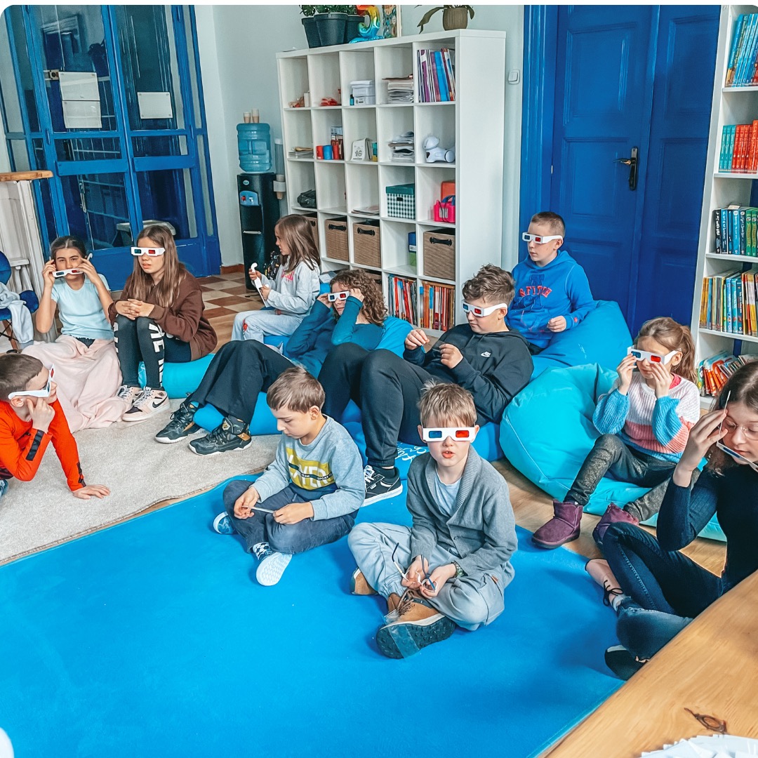 Grupa dzieci w okularach 3D siedzi na podłodze i pufach w jasnym pomieszczeniu z regałami na książki.