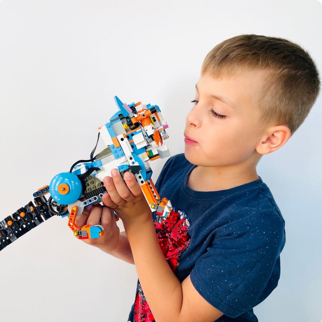 Chłopiec z zamkniętymi oczami trzyma w rękach konstrukcję z klocków Lego, przypominającą futurystyczną broń z elementami pomarańczowymi, białymi i niebieskimi.