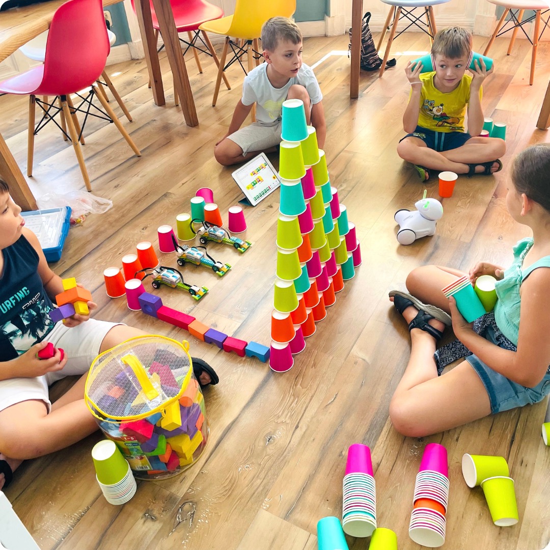Dzieci siedzą na podłodze bawiąc się kolorowymi kubkami, klockami i robotem edukacyjnym; w tle widoczne krzesła w różnych kolorach.