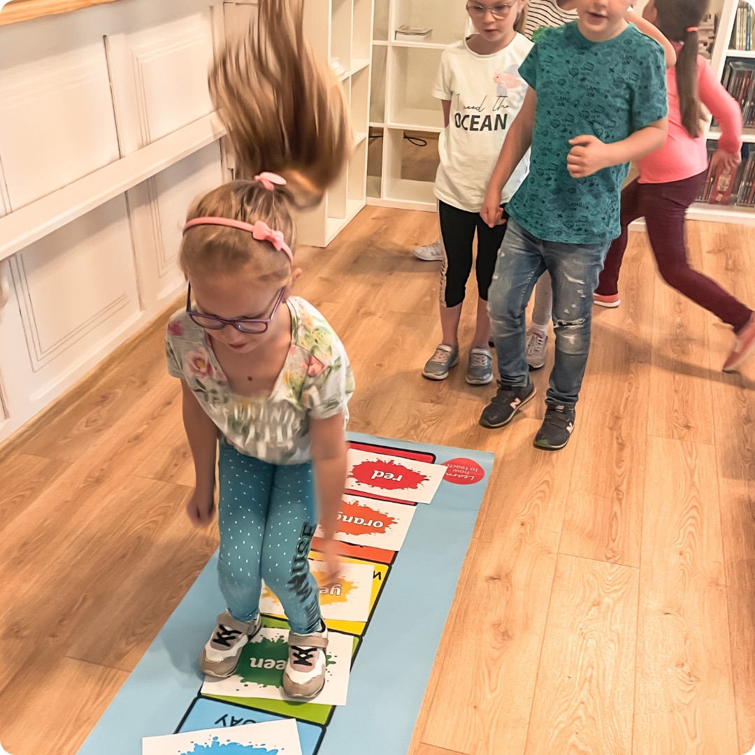 Dziewczynka w okularach skacze na macie z kolorami podczas zajęć grupowych, widoczni inni uczniowie i regał z książkami w tle.