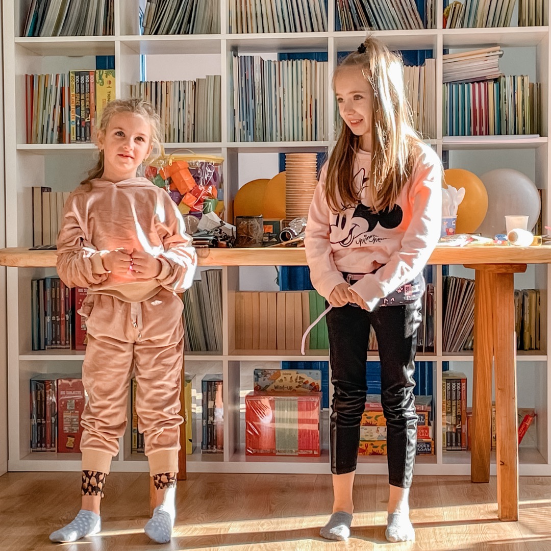 Dwie dziewczynki w jasnych ubraniach stoją przed regałem z książkami i zabawkami, prawdopodobnie po lekcji angielskiego online, w słoneczny dzień.