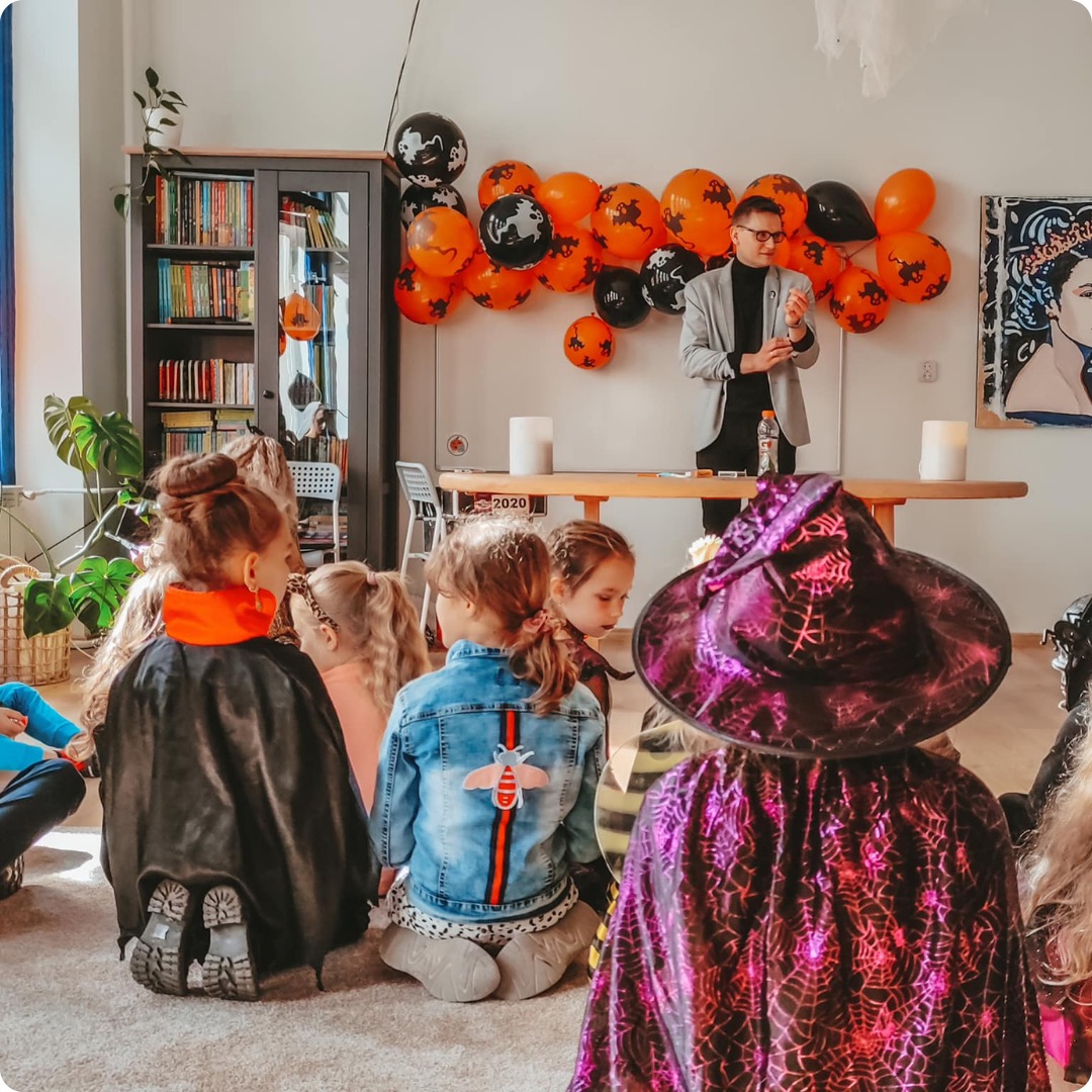 Mężczyzna w szarej marynarce prezentuje coś dzieciom w kostiumach na tle pomarańczowo-czarnych balonów i regału z książkami.