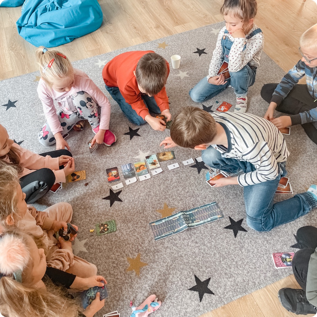 Grupa dzieci siedzi na dywanie w gwiazdki, grając w grę karcianą z ilustracjami i numerami, z niebieskim pufem w tle.