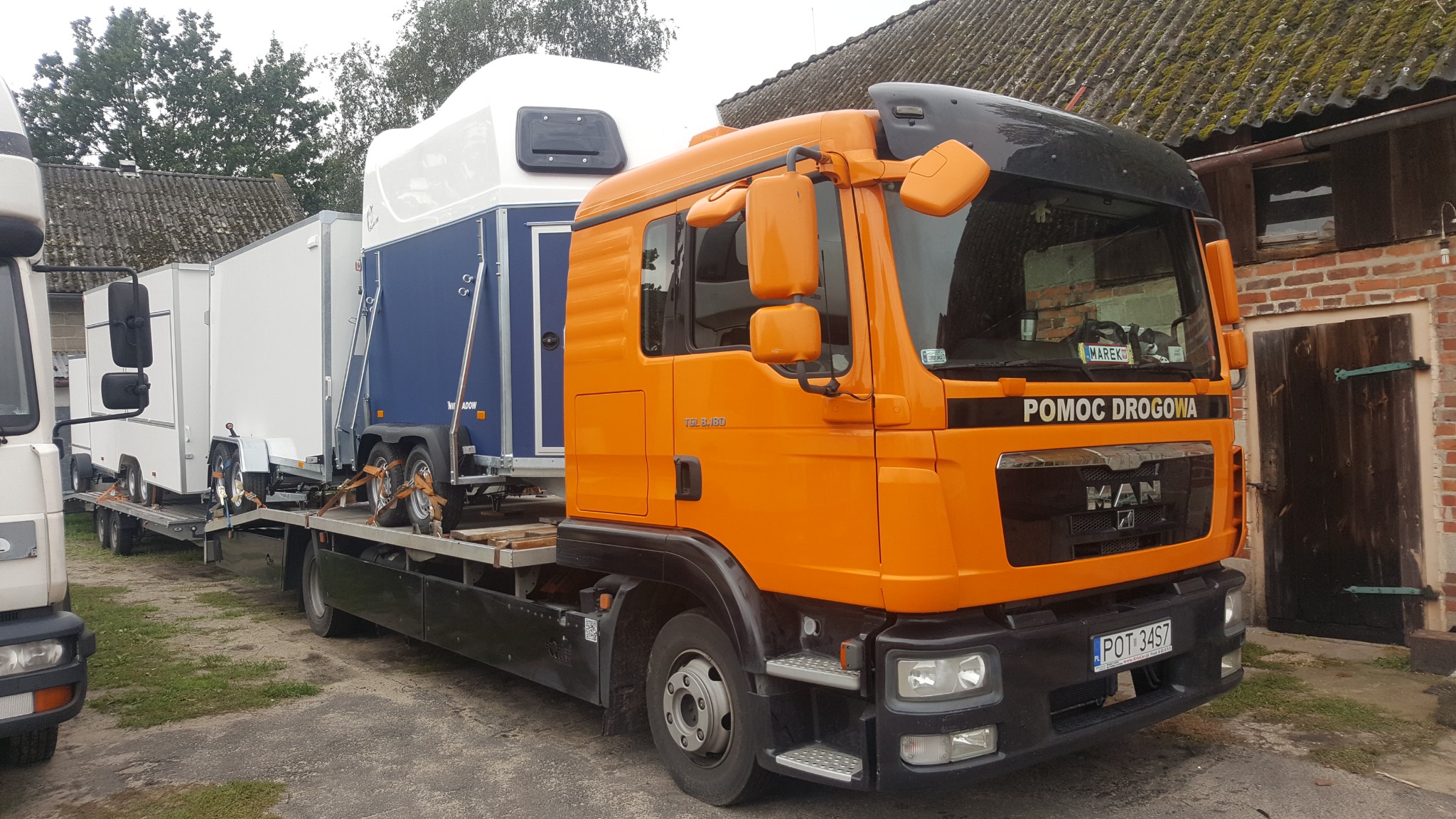 Pomarańczowa laweta MAN z przyczepą przewożącą dwie biało-niebieskie przyczepy kempingowe, na tle ceglanego budynku z drewnianymi drzwiami.