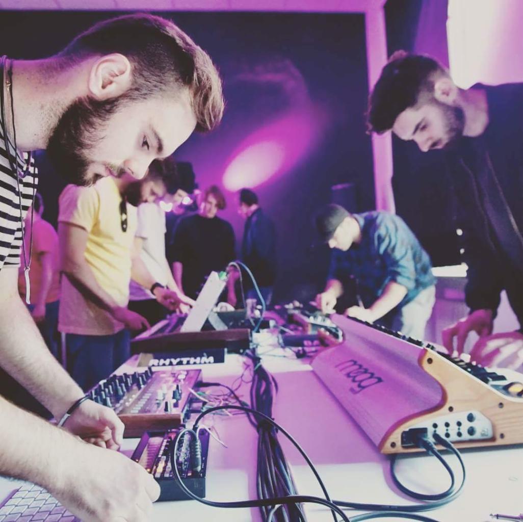 Grupa muzyków grająca na syntezatorach Moog i innych instrumentach elektronicznych w fioletowym oświetleniu scenicznym, widoczne kable i miksery.