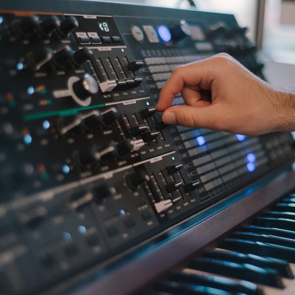 Ręka reguluje suwak na panelu syntezatora z podświetlanymi przyciskami i klawiaturą w tle.