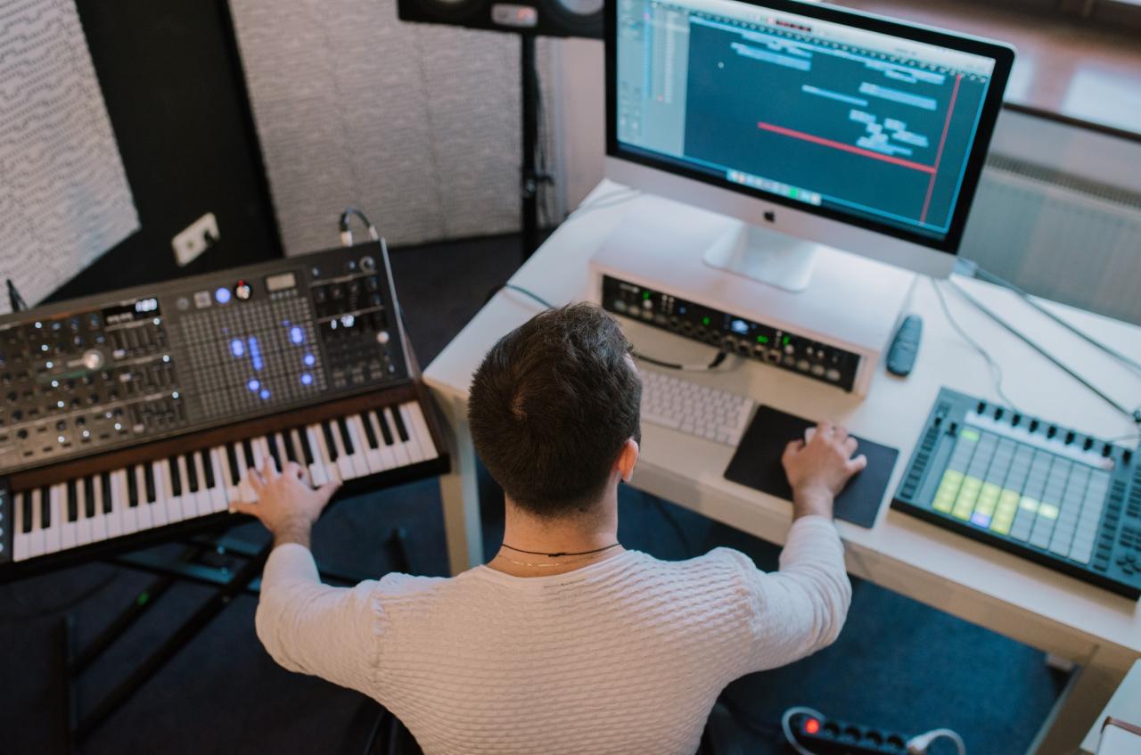 Mężczyzna od tyłu pracuje przy biurku z syntezatorem, klawiaturą MIDI, mikserem audio i komputerem z oprogramowaniem do edycji dźwięku.