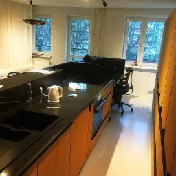 Nowoczesna kuchnia z czarnym blatem i drewnianymi frontami, widok na zlew, czajnik elektryczny i okno z widokiem na drzewa.