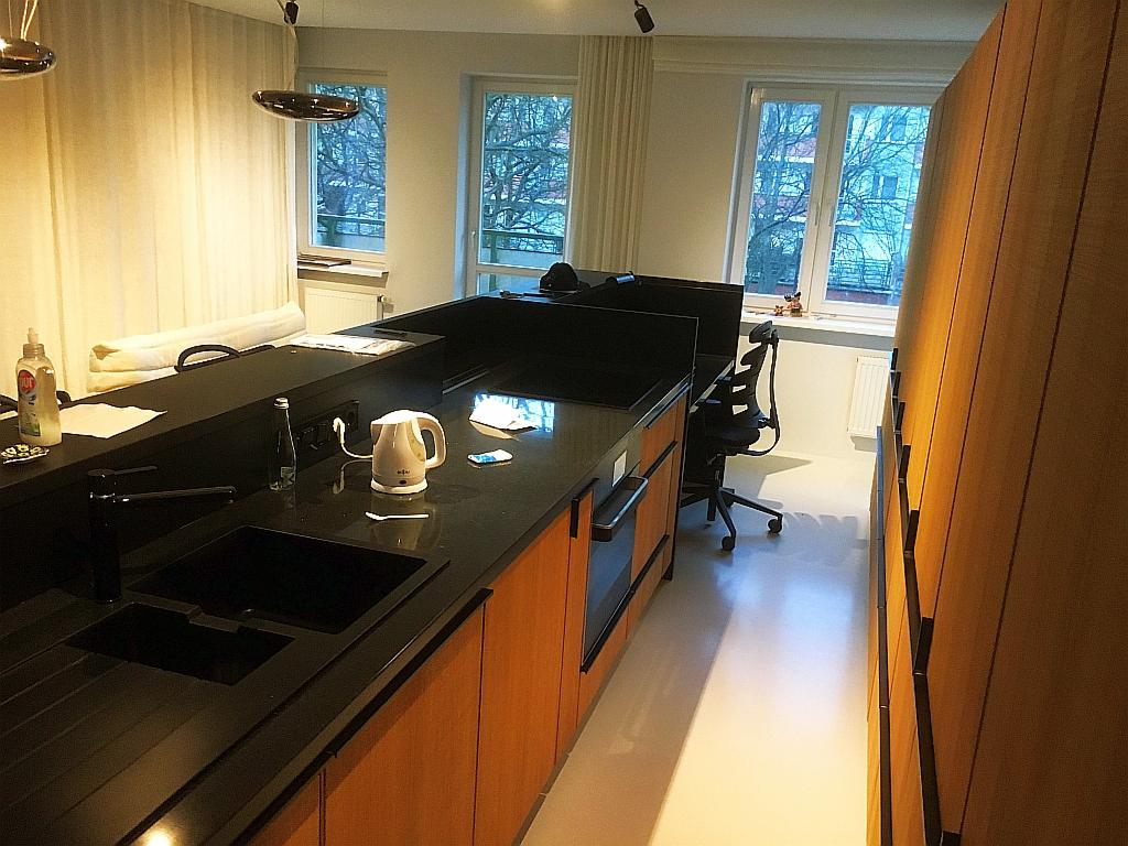 Nowoczesna kuchnia z czarnym blatem i drewnianymi frontami, widok na zlew, czajnik elektryczny i okno z widokiem na drzewa.