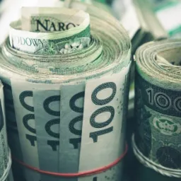 Zwinięte w rulony banknoty 100 zł zabezpieczone gumkami recepturkami i sznurkiem, ułożone w stosy.