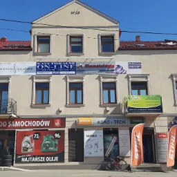 Fasada budynku z kilkoma firmami: części samochodowe, serwis laptopów, kasy fiskalne, stomatologia, fizjoterapia, agro-tech, widoczne szyldy i reklamy.