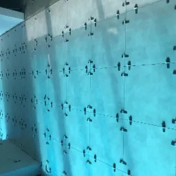 Ściana z szarych płyt przygotowywana do położenia płytek, widoczne krzyżyki dystansowe i sznurki poziomujące, oświetlenie w odcieniach błękitu.