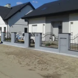 Nowoczesne domy jednorodzinne z szarą elewacją, czarną dach&oacute;wką i ozdobnym, metalowym ogrodzeniem pomalowanym na czarno, z numerem domu '22' na słupku.