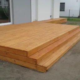 Drewniany taras z schodami, świeżo po montażu w Gąsawach, przylegający do budynku z jasną elewacją i roletami zewnętrznymi.