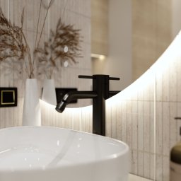 Mudi Agnieszka Kasprzyk - Nowoczesna łazienka z owalną umywalką, czarną armaturą i podświetlanym lustrem. Beżowe płytki imitujące kamień i dekoracyjne trawy pampasowe dodają elegancji.