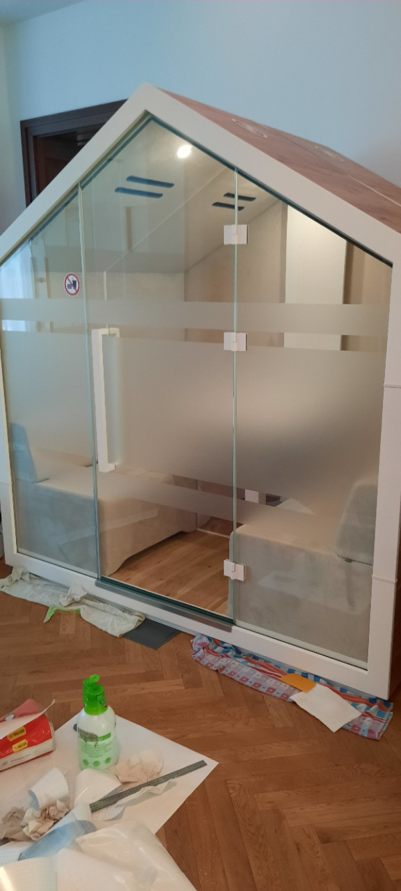 Aplikacja folii matowej na szklanej konstrukcji w kształcie domku, widoczne narzędzia pracy. Usługa wykonana w Szczecinie, częściowo przyciemnione szyby.