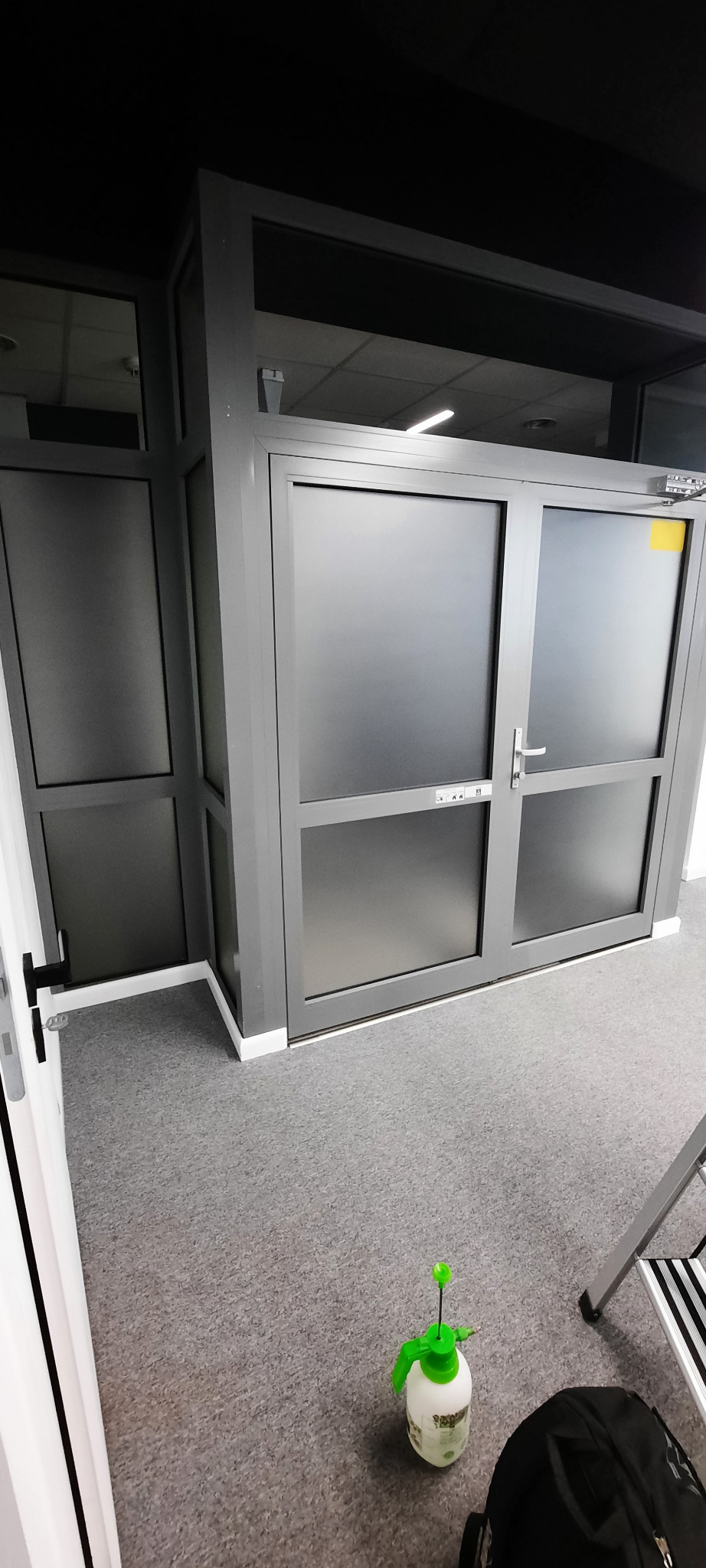 Szare, przeszklone drzwi z nałożoną folią, widoczne akcesoria montażowe, butelka ze spryskiwaczem i fragment drabiny, szara wykładzina podłogowa.