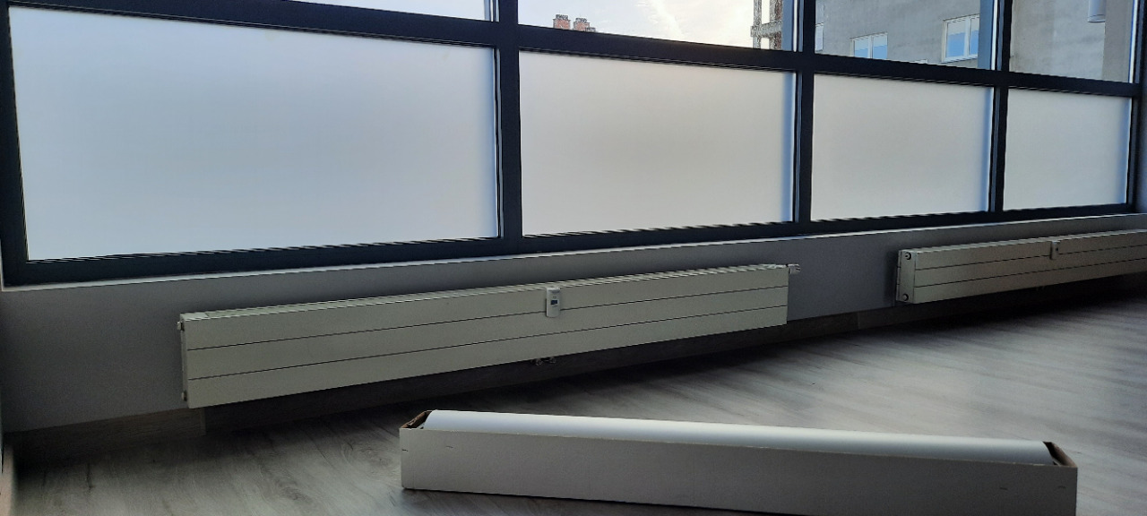 Okna biurowe z folią matującą, widoczne grzejniki i pudełko z folią. Matowe szyby zapewniają prywatność w nowoczesnym wnętrzu biura.