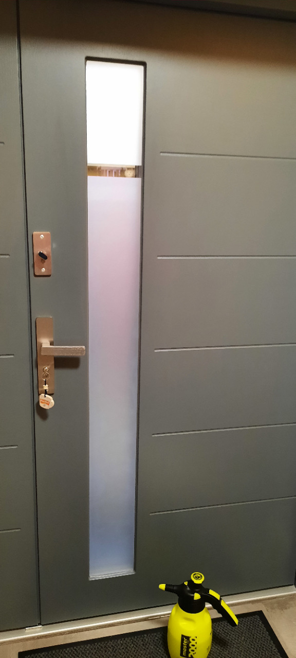Szare drzwi wejściowe z pionowym, częściowo przyciemnionym szybem. Na wycieraczce stoi żółty spryskiwacz. Widoczne detale: klamka, zamek i klucze.